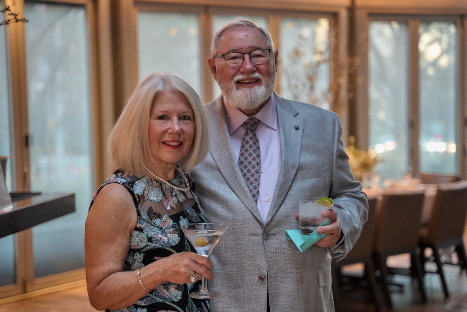 Two guests smiling together holding cocktails at an indoor evening gathering near a set dinner table