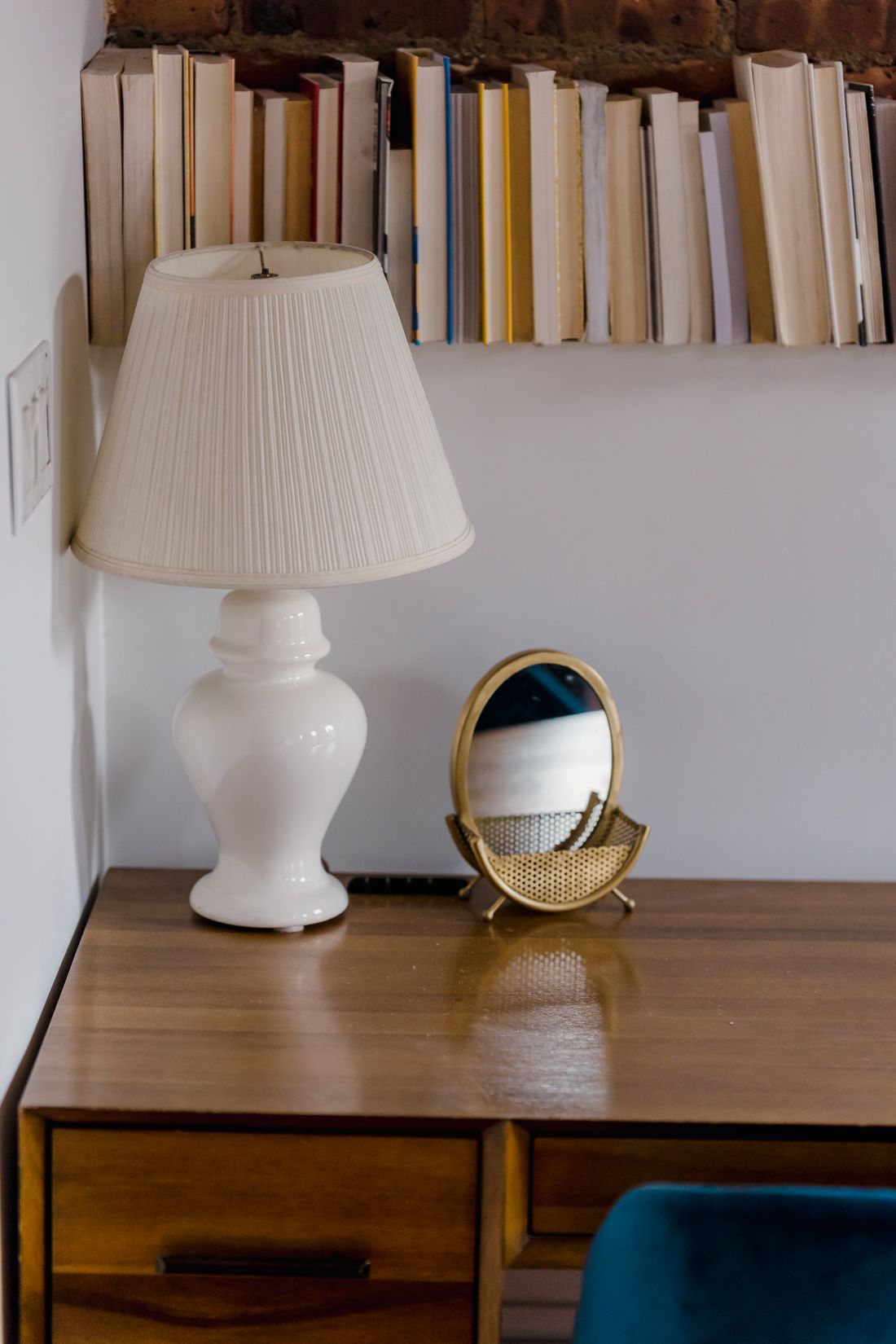 A wooden desk with a table lamp and a small mirror, with a row of books on a shelf above lit by daylight.