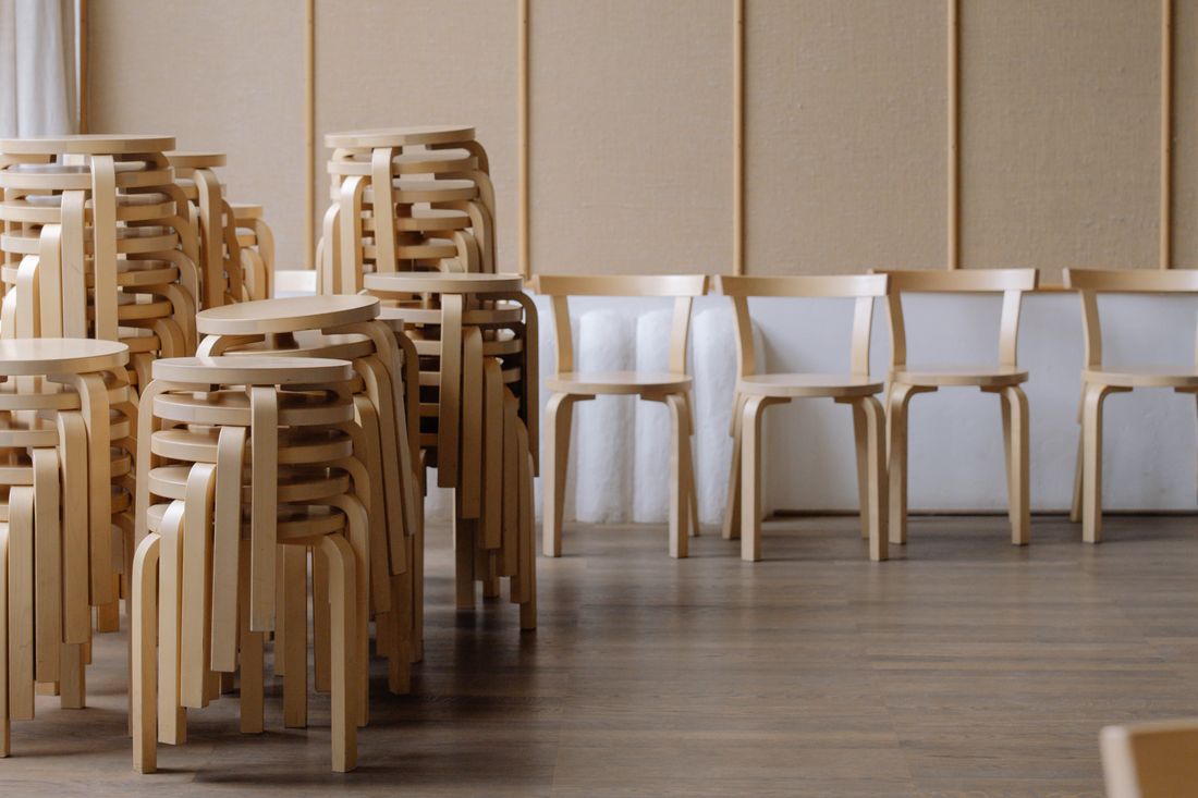 Stacked wooden stools next to a row of matching wooden chairs set up along a padded wall in a plain room with wood flooring, no people visible.