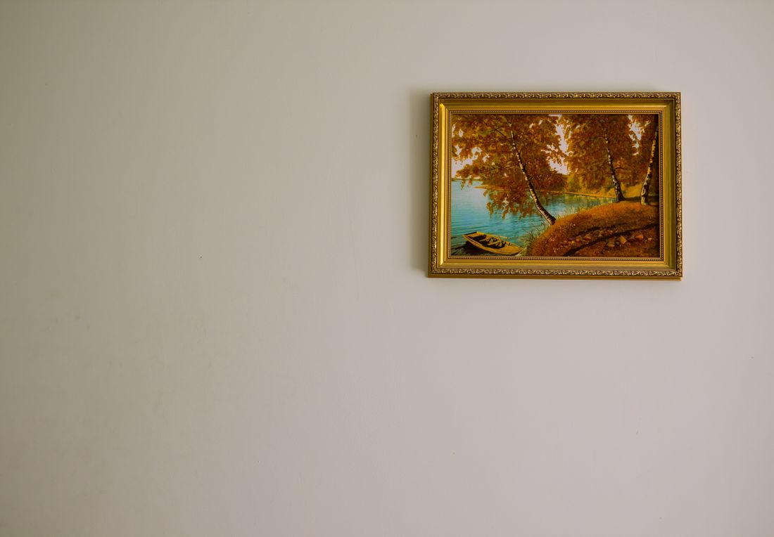 A single gold-framed landscape painting hangs on a plain light-colored wall with open space around it.