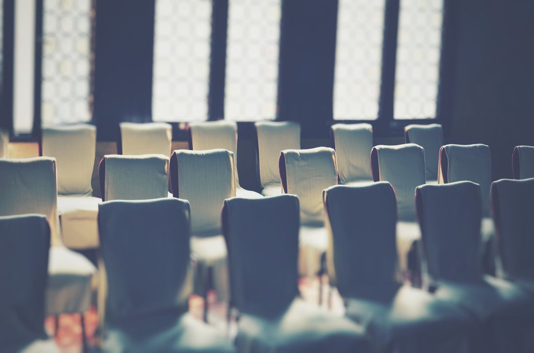 Rows of empty fabric-covered chairs arranged in a plain room lit by tall windows, set up ahead of a gathering