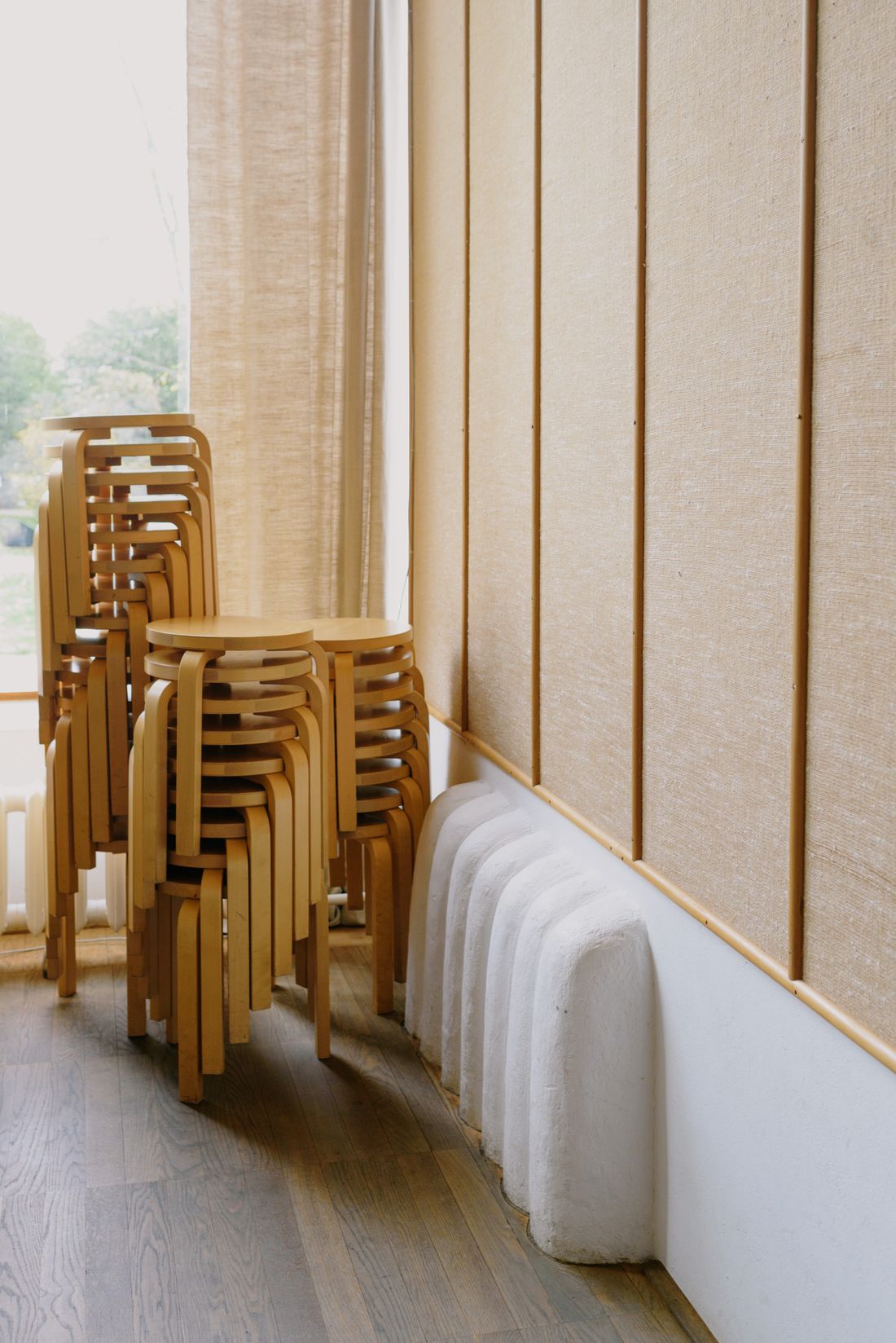 Stacks of wooden stacking chairs pushed into the corner of a plain room next to a window