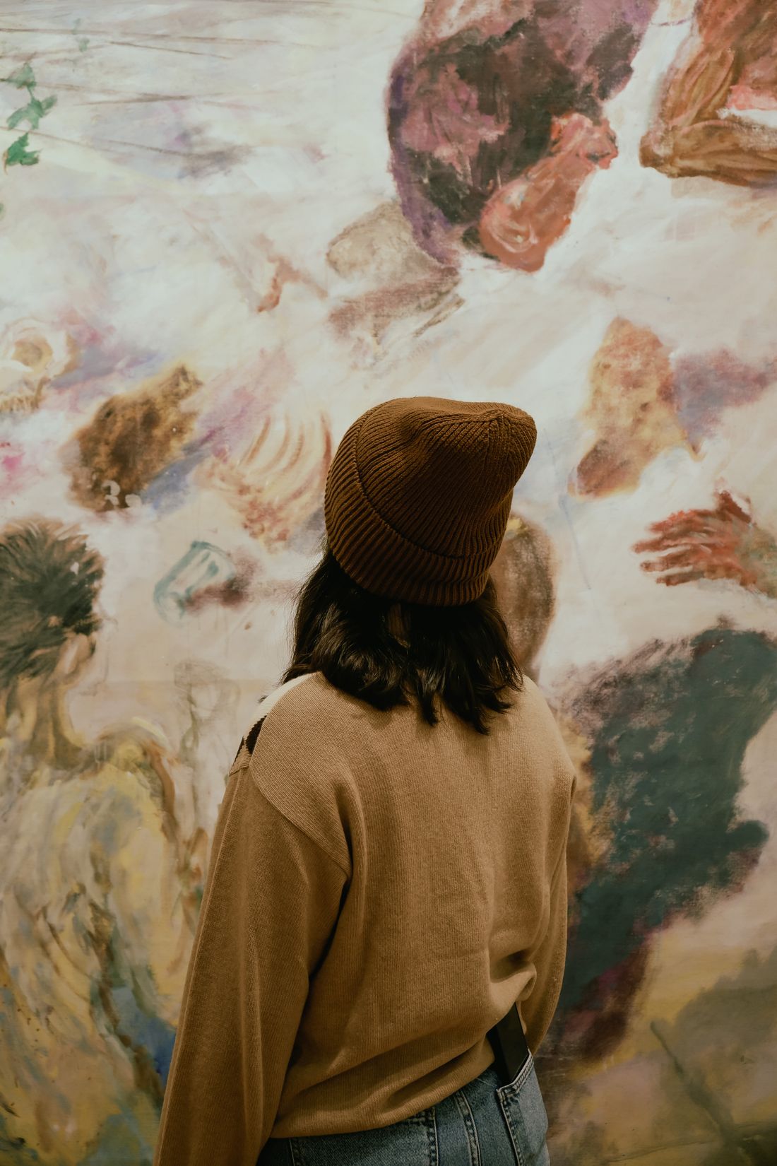 A person in a beanie viewed from behind, standing close to a large abstract painting in a gallery.