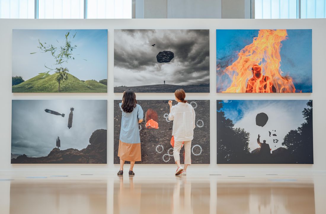 Two visitors seen from behind standing close together in a bright white gallery, looking up at large photographic works spanning the wall under a row of high windows.