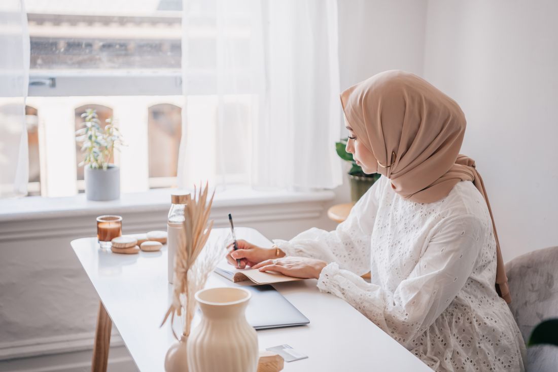 A woman in a hijab seated at a plain desk by a window, writing in a notebook, side view.