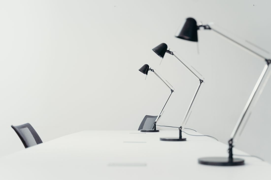 Three black desk lamps lined up on an empty white desk with empty chairs, plain wall behind, no people.