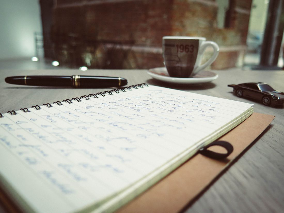 A handwritten notebook, fountain pen, and coffee cup on a wooden table