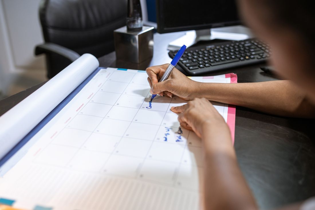 A hand marking dates on a paper desk calendar with a pen near a keyboard
