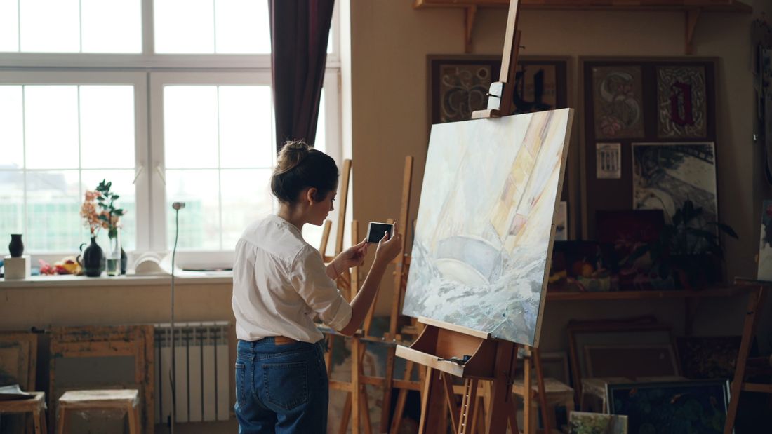 A person photographing a landscape painting on an easel with a phone in a studio