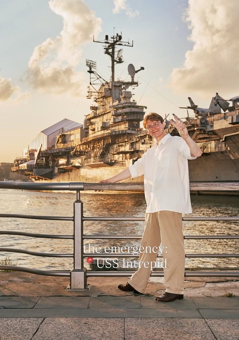 Ian Sowden leaning on a Hudson River railing at sunset throwing a peace sign, with the USS Intrepid and its aircraft behind him.