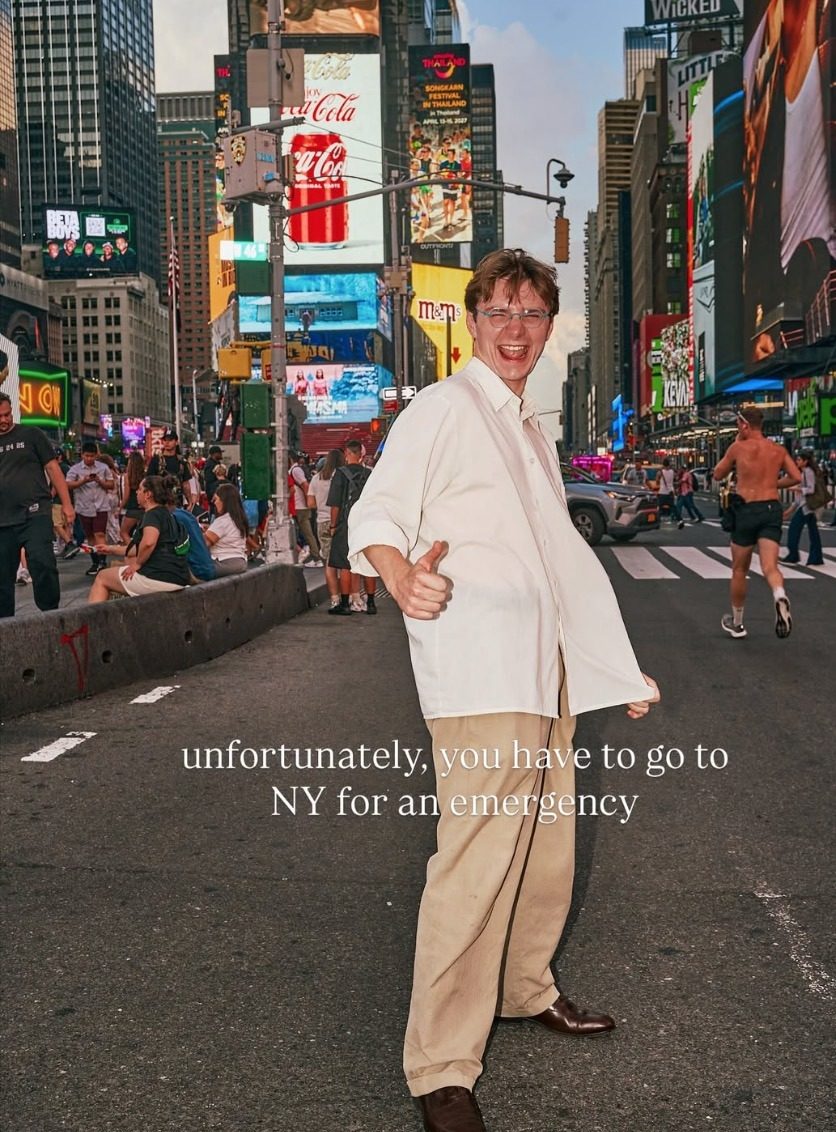 Ian Sowden giving a thumbs up mid-stride in a Times Square crosswalk at dusk, wearing an oversized white shirt and tan trousers.