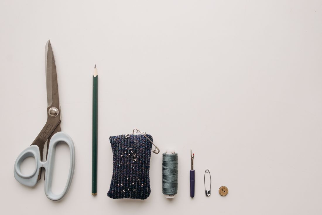 A flat lay of basic styling and sewing tools, including scissors, a pencil, thread, safety pins, and a button, on a plain background