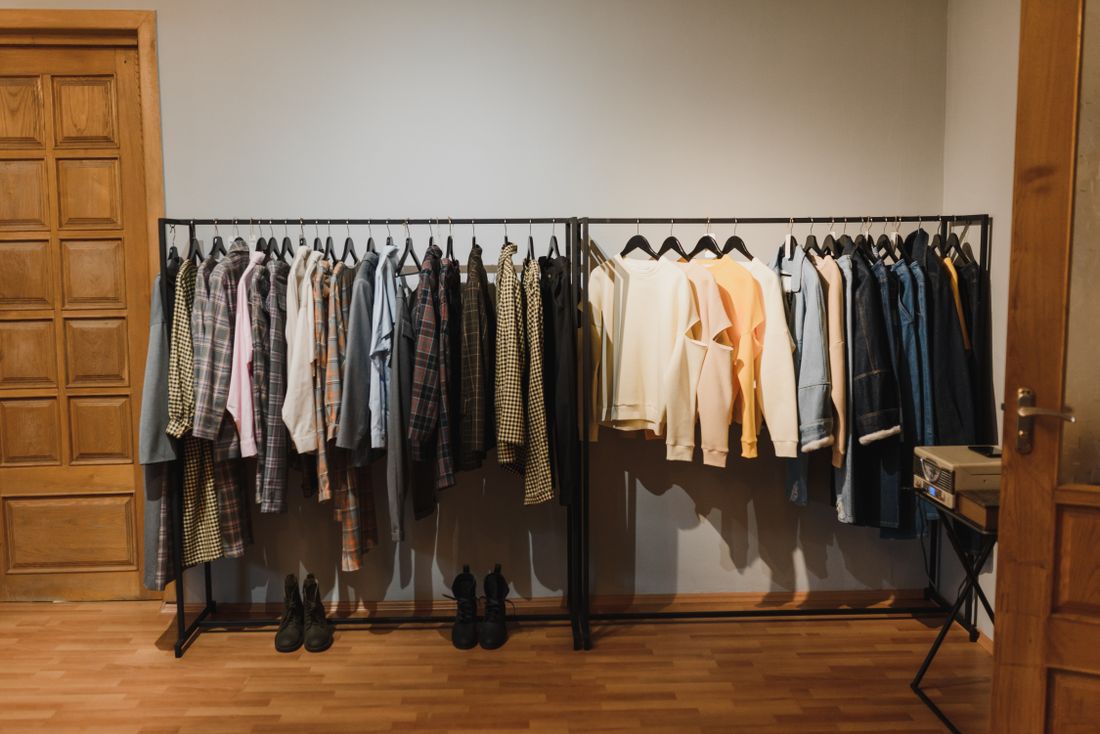 Two clothing racks holding a wide range of shirts, jackets, and pants sorted by tone in a bright room with wood flooring