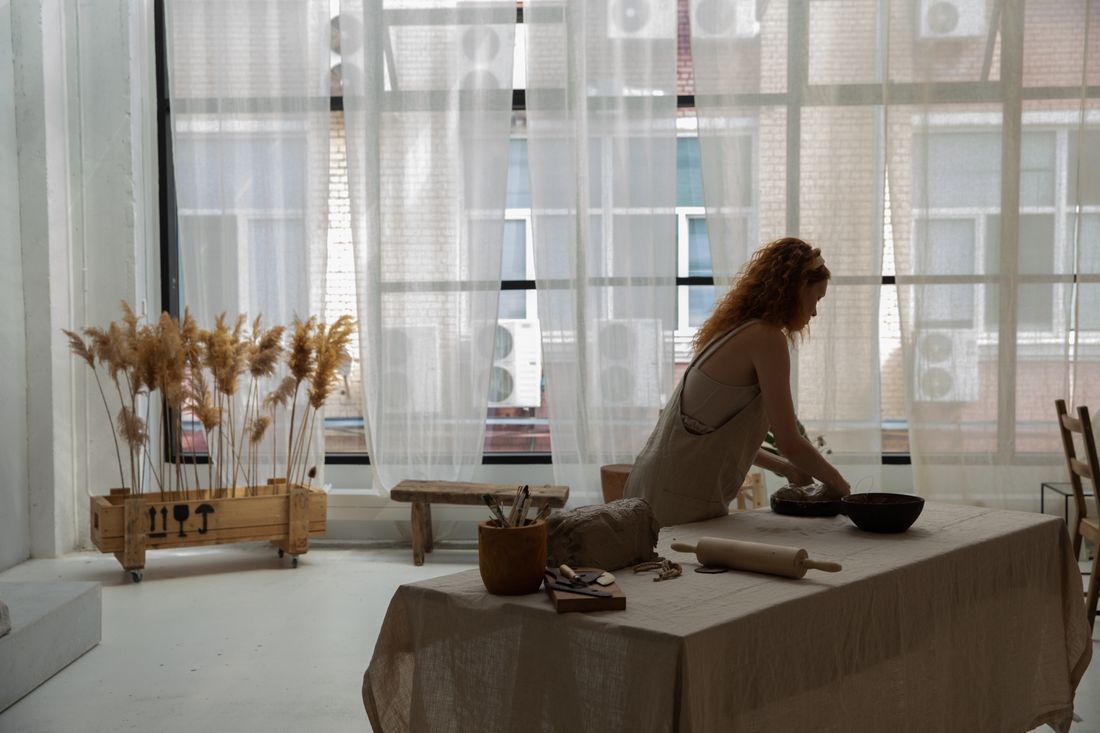 A person in a plain work smock sits on a cloth-covered table shaping clay by a wall of tall studio windows, face turned away.