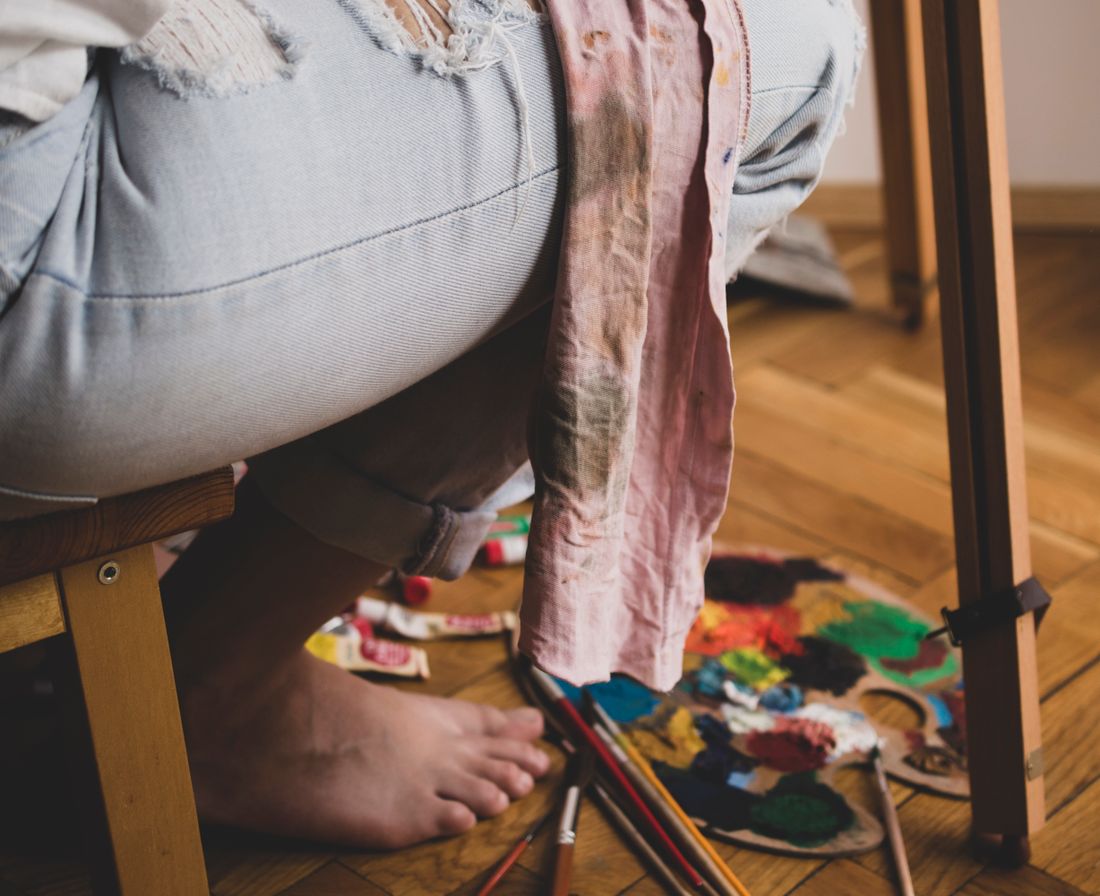 Close view of light denim jeans and a paint-marked cloth hanging from a wooden stool, with a used paint palette and brushes on the floor below.