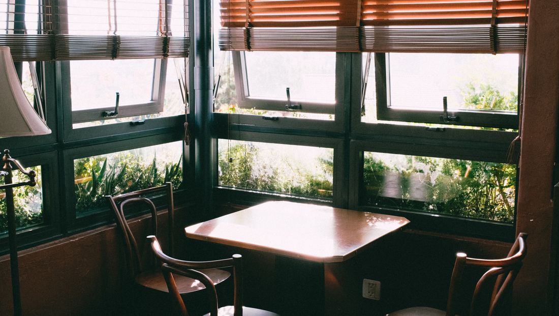 A quiet corner room with two wooden chairs at a small table, daylight coming through half-drawn blinds and greenery outside the windows.