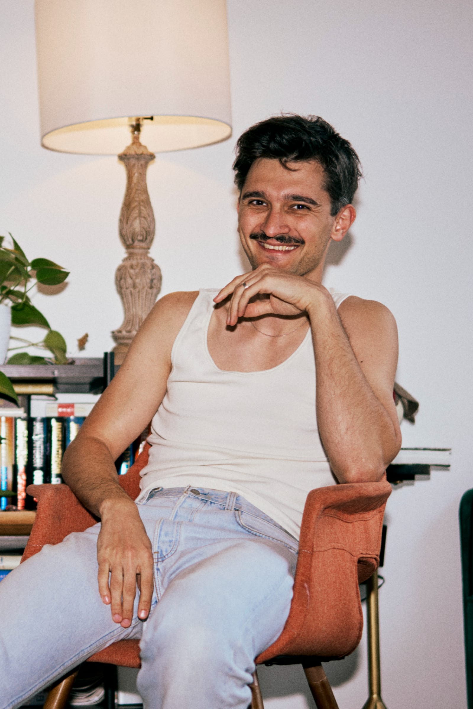 A man in a white tank top sits smiling in an orange armchair beside a lamp and a bookshelf in a lived-in room.
