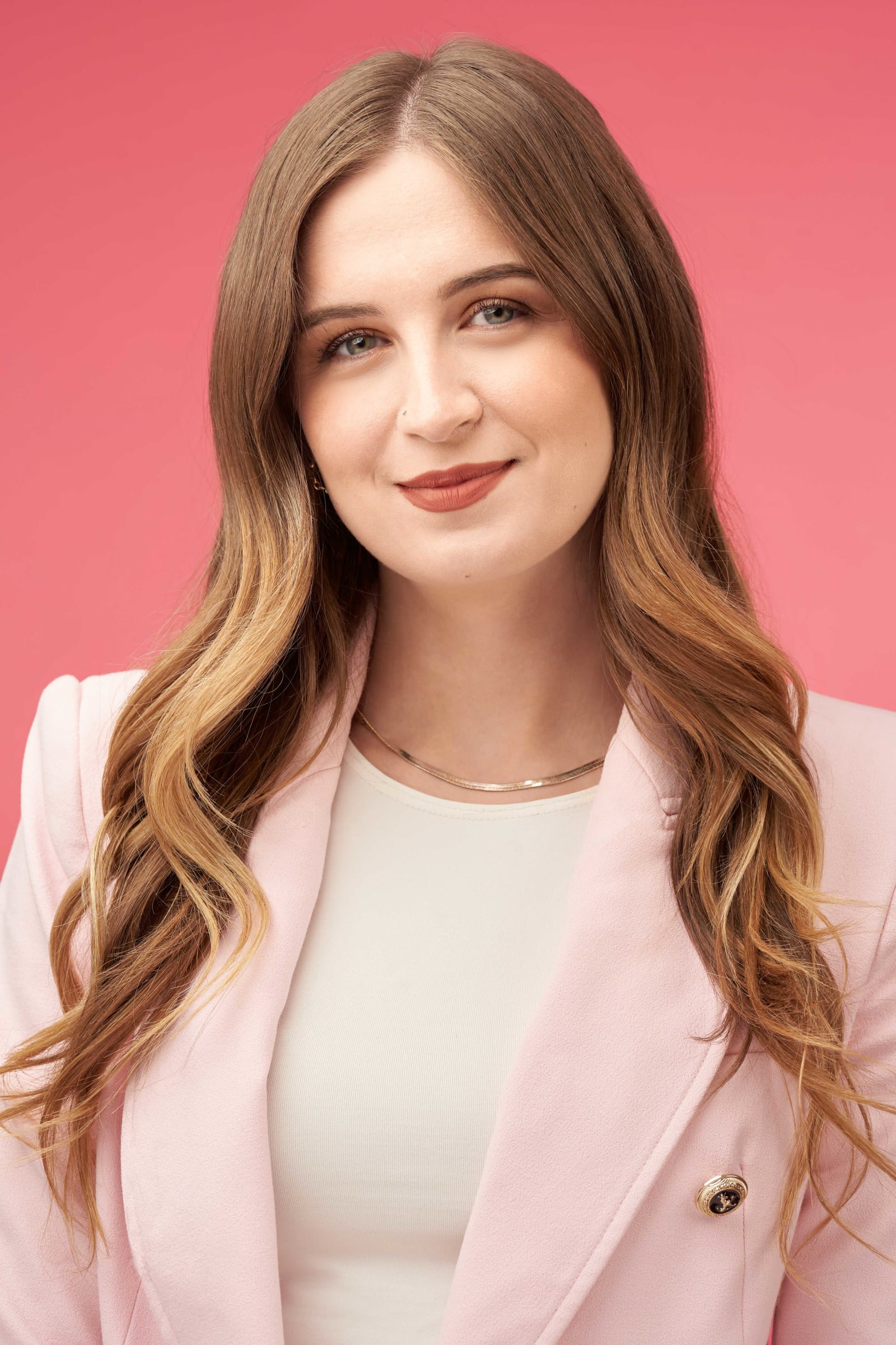 A smiling young woman in a pink blazer photographed against a plain coral pink studio backdrop.