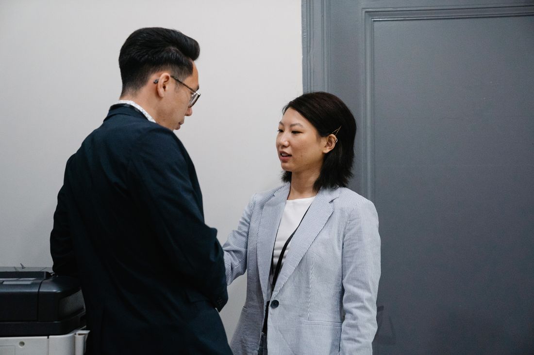Two adults in conversation standing close together indoors against a plain wall.