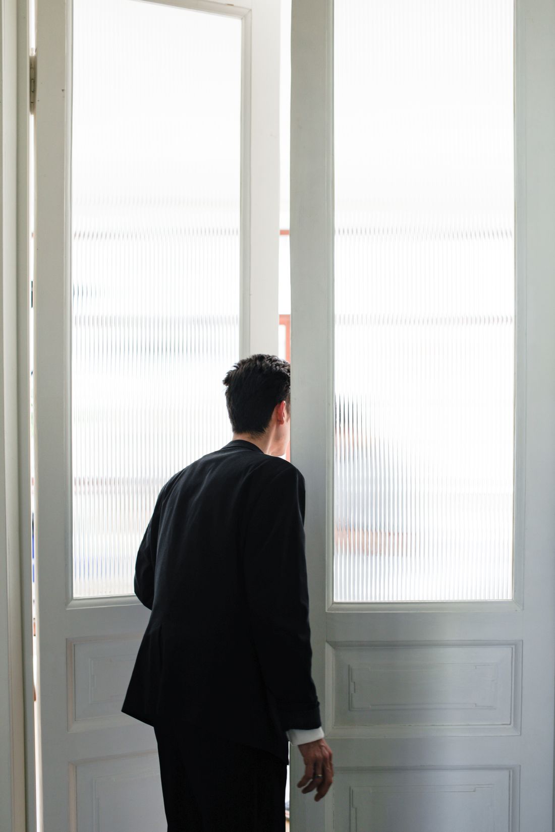 A person seen from behind pausing at a plain interior doorway, looking through the glass.