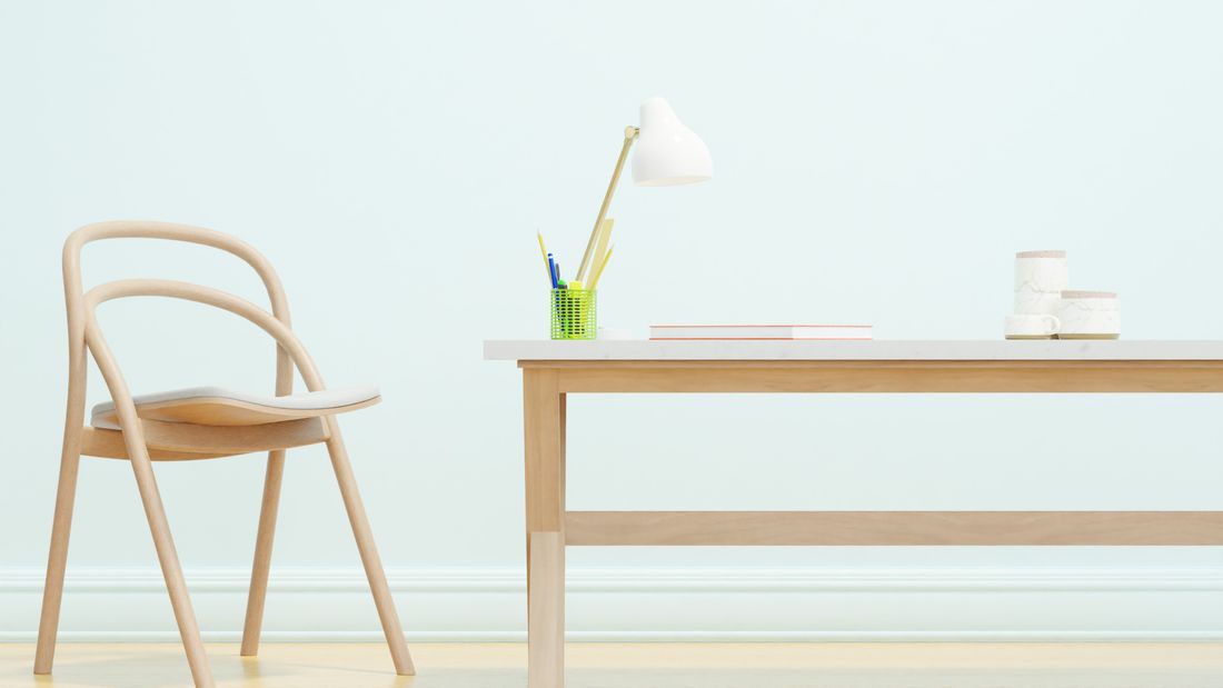 A plain desk and chair in a simple room, with a notebook and a couple of mugs on the desktop and no one in frame.