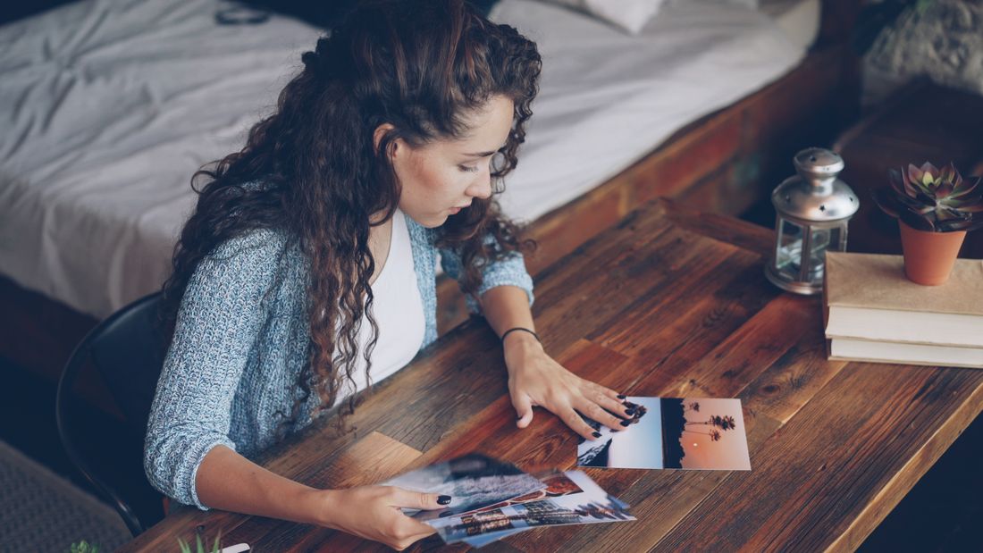 A person seated at a wooden desk, holding printed photographs and looking down at more prints laid across the table.