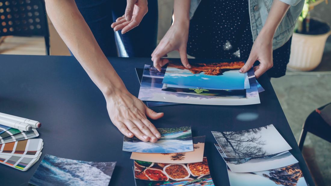 Two people's hands and arms over a dark table sorting printed landscape photographs, with a color swatch booklet nearby.