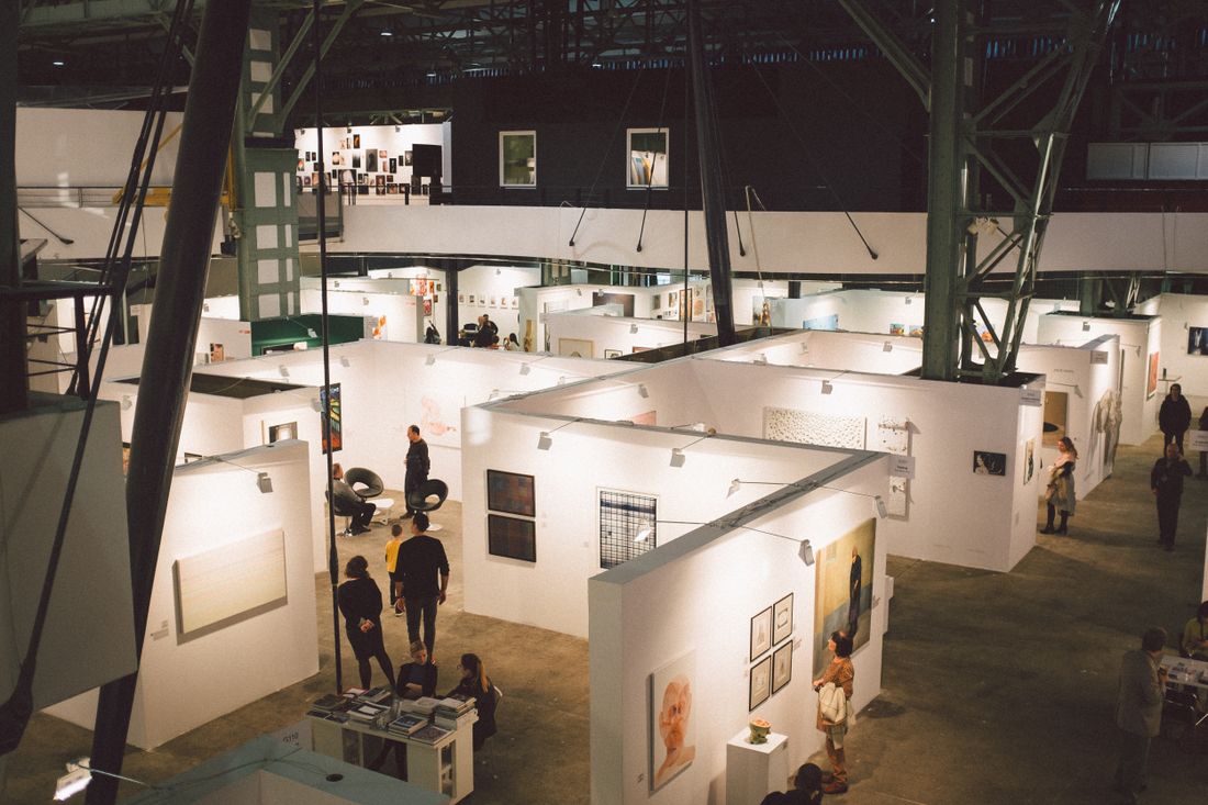Wide aerial view of an indoor art fair with framed artwork on temporary booth walls and visitors browsing in the distance.