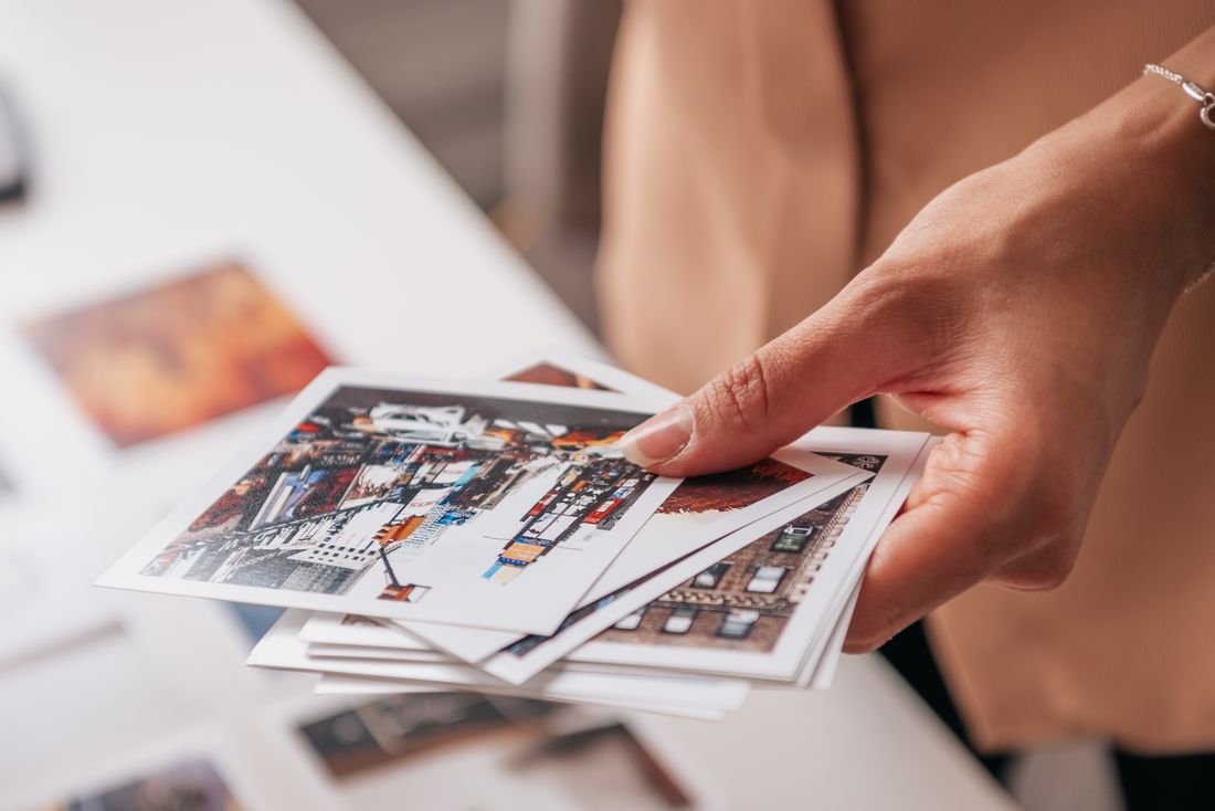 Close angled view of a hand holding a fanned stack of printed photographs, with more prints softly out of focus on the table beyond.
