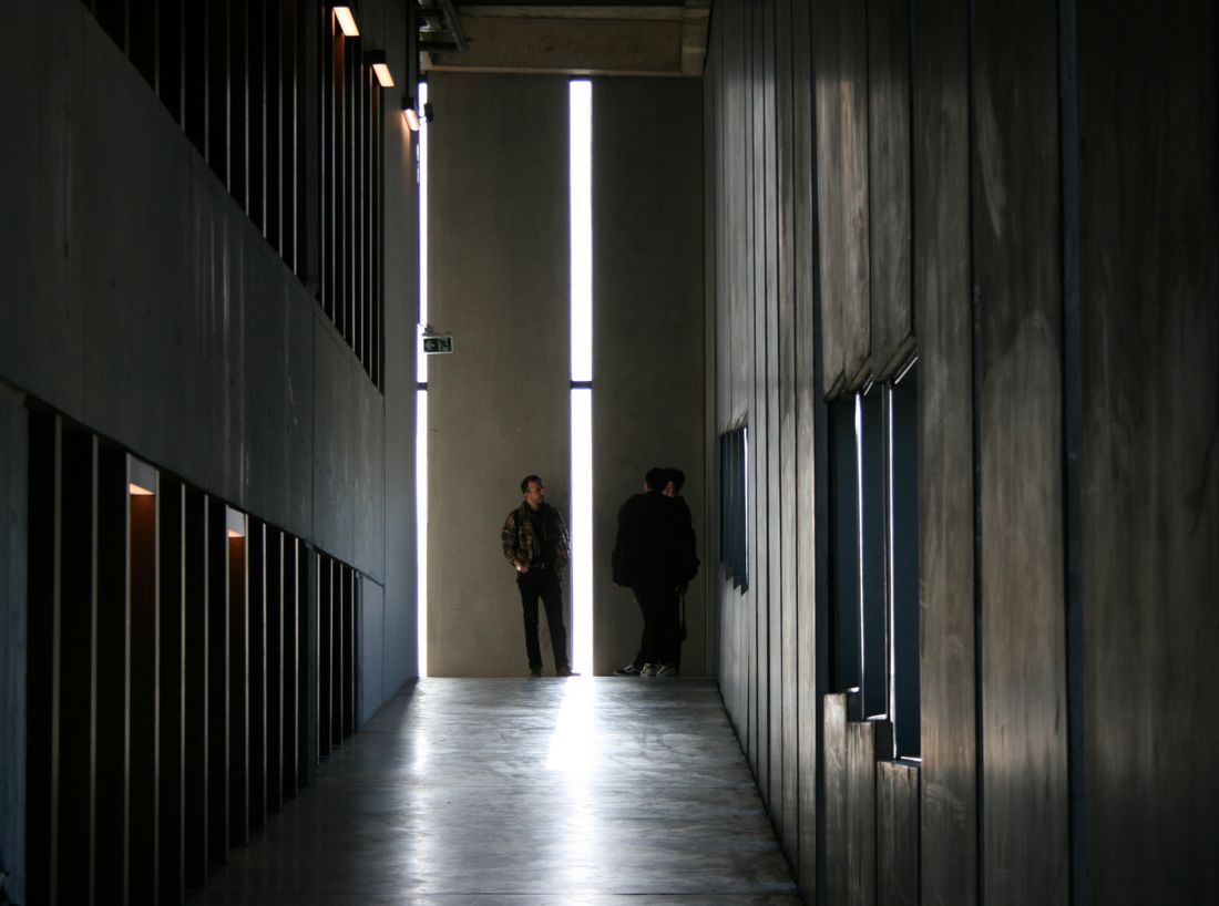 Two people talking at a distance inside a dim, plain concrete corridor with wood-paneled walls.