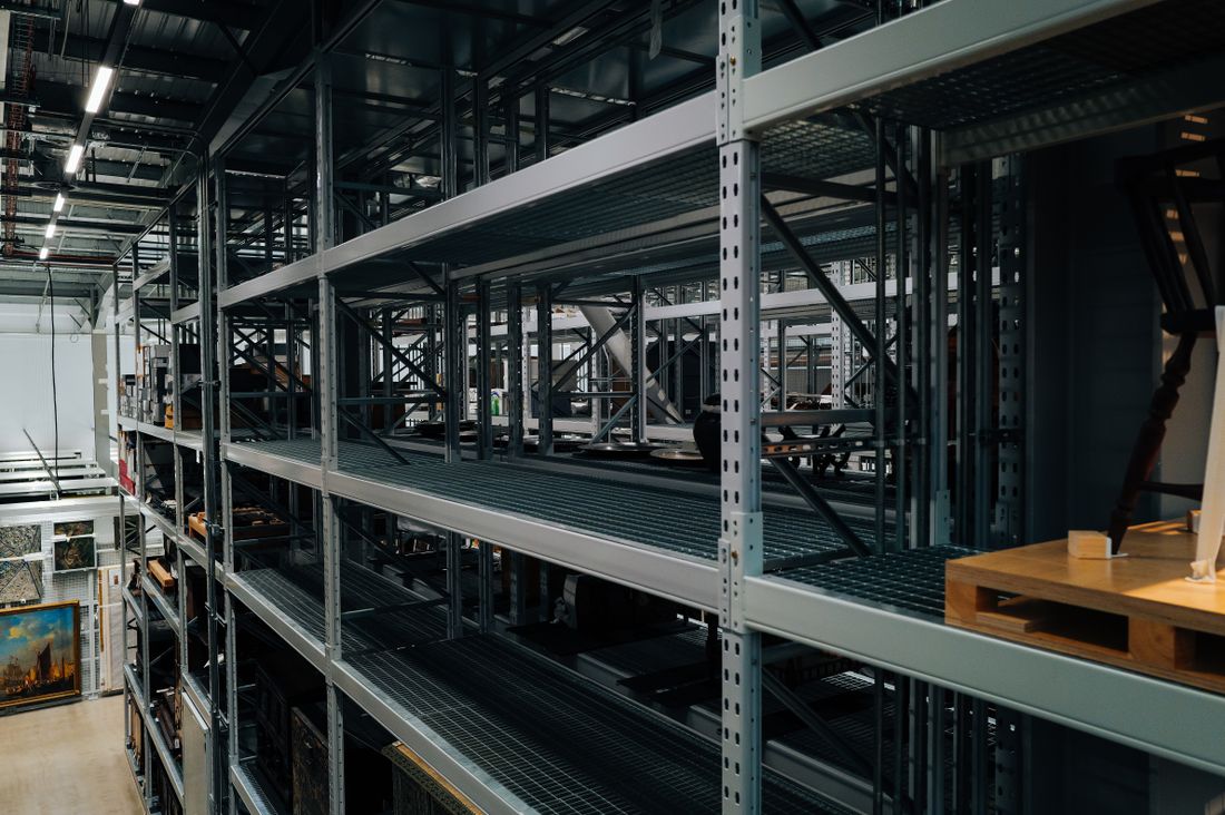 Tall metal storage racking in an industrial room, with framed paintings stored upright along the floor.