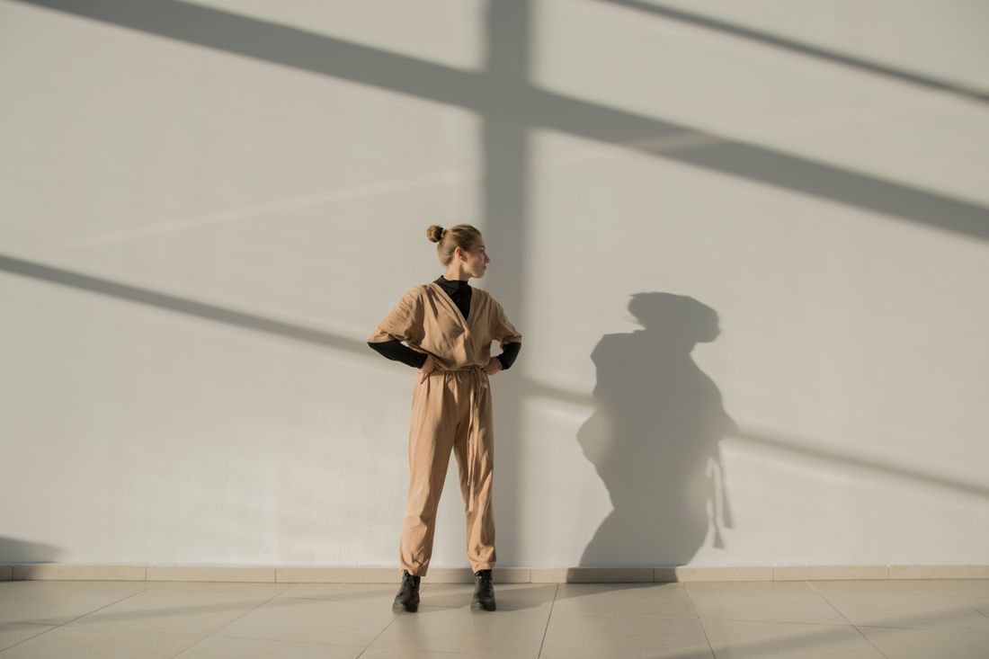 A person in a plain tan jumpsuit and black boots stands at a distance against a bright white wall crossed by window-light shadows