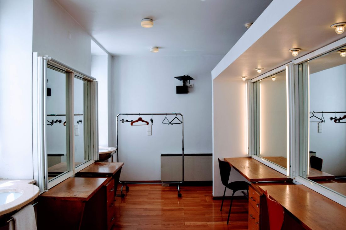 An empty dressing room with mirrored counters, a wooden floor, and a rolling rail holding a few bare hangers.