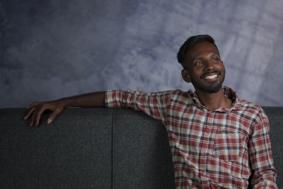 A man seated indoors, smiling naturally, wearing a plaid flannel shirt against a plain studio backdrop.