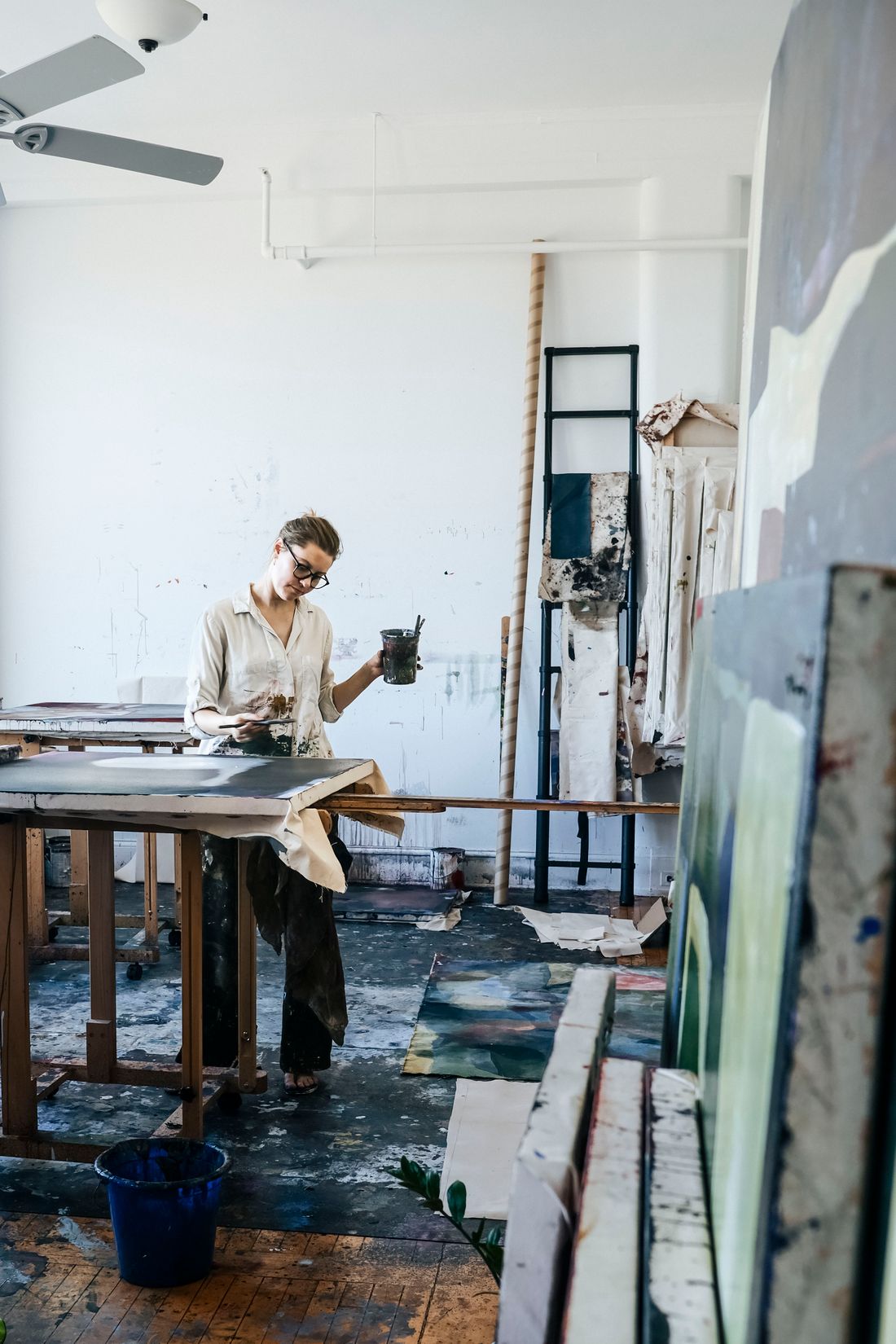 A woman artist standing in her working studio, holding a paint-mixing cup, surrounded by work-in-progress canvases and materials.