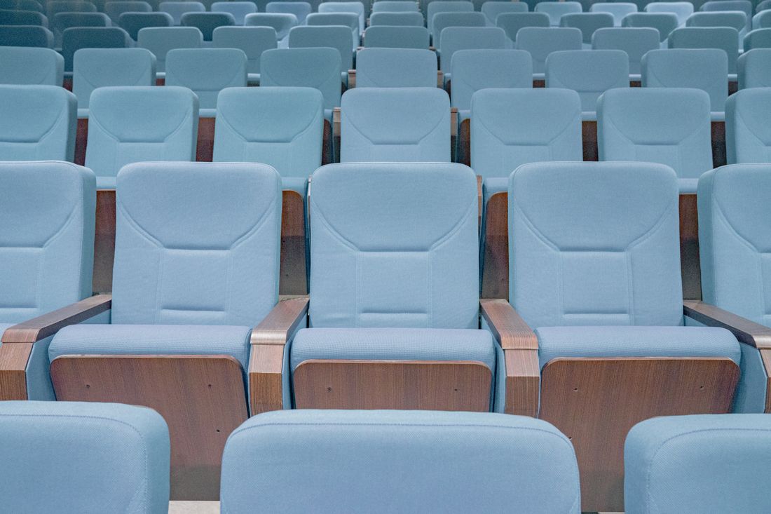 Rows of empty upholstered auditorium seats facing forward, arranged in tight rows with no audience present.