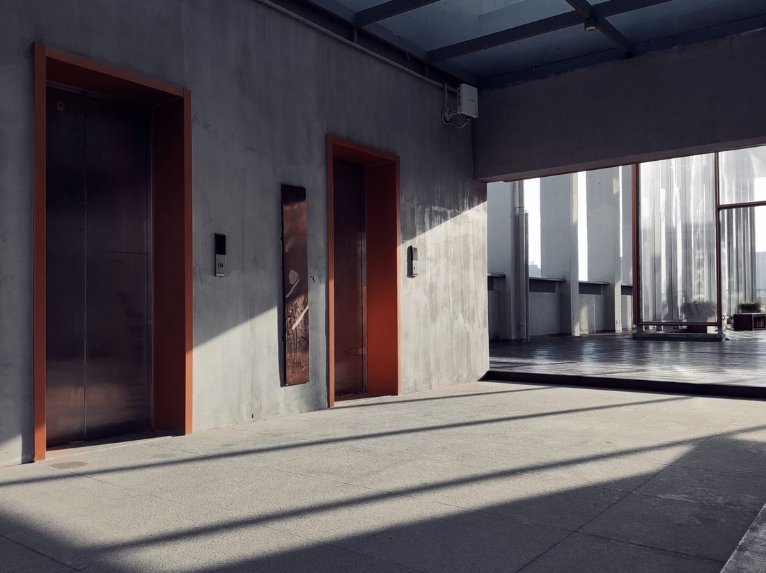 An empty concrete interior with two elevator doors, sharp afternoon light and shadow crossing the floor toward an open terrace.