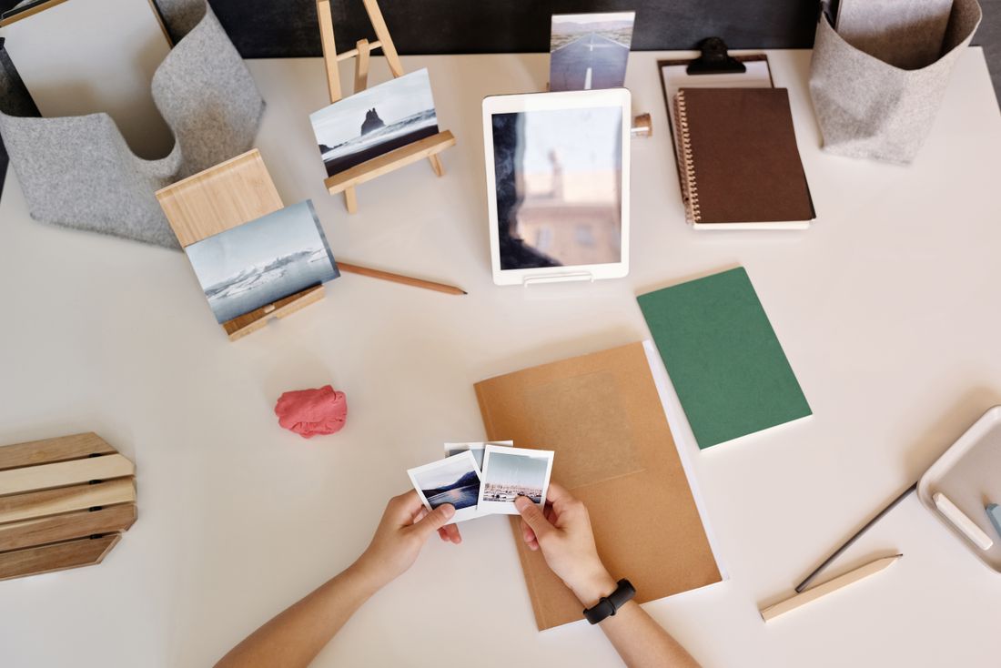 Overhead view of two hands holding small printed photographs above a desk, with more prints displayed on small wooden stands and a tablet nearby.