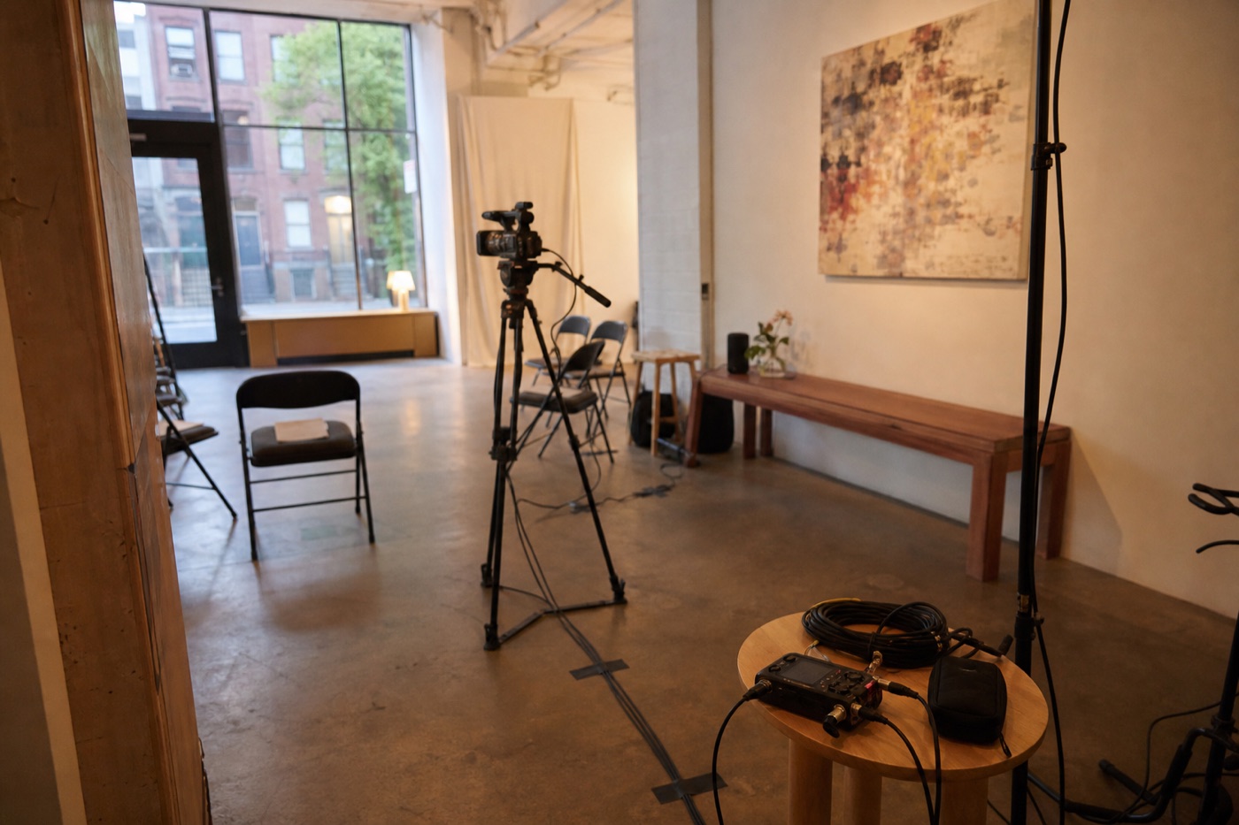 Small creative event room before guests arrive with a tripod camera, taped cables, chairs, artwork, and an audio recorder set up at a distance