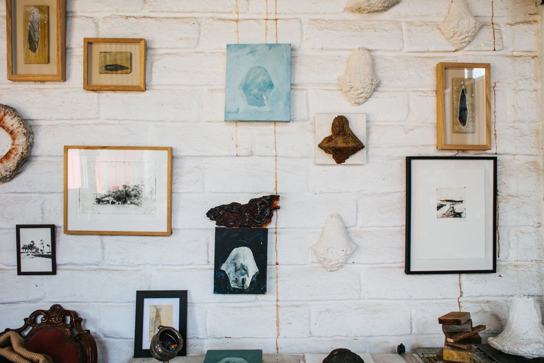 A white brick wall hung with framed prints, small unframed panels, and plaster wall objects in a range of sizes, media, and frame styles.