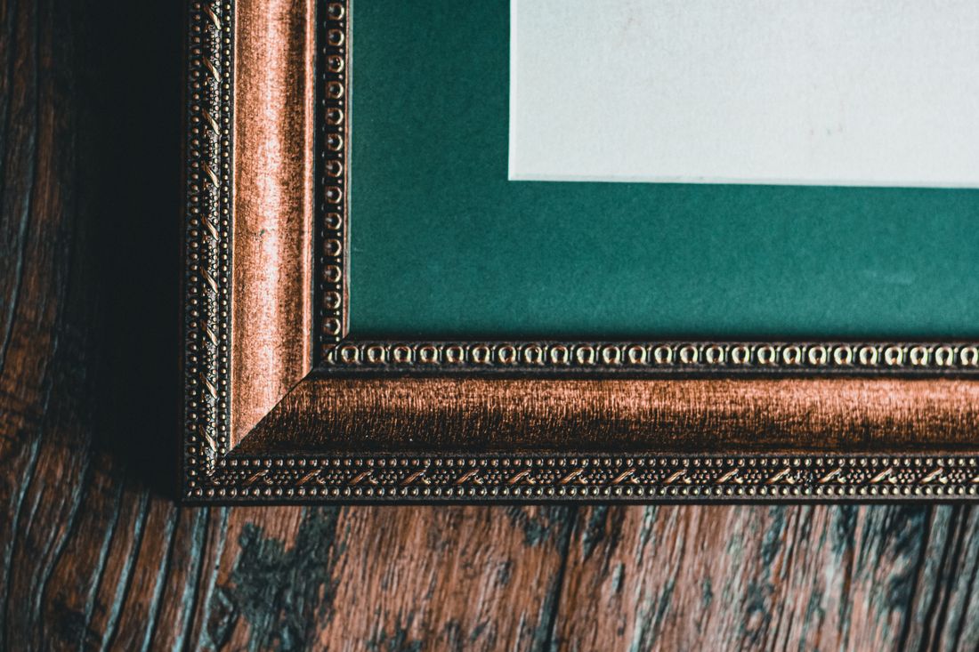 A close view of an ornate gilt frame corner with a deep green mat around a blank white sheet, resting on a dark wood surface.