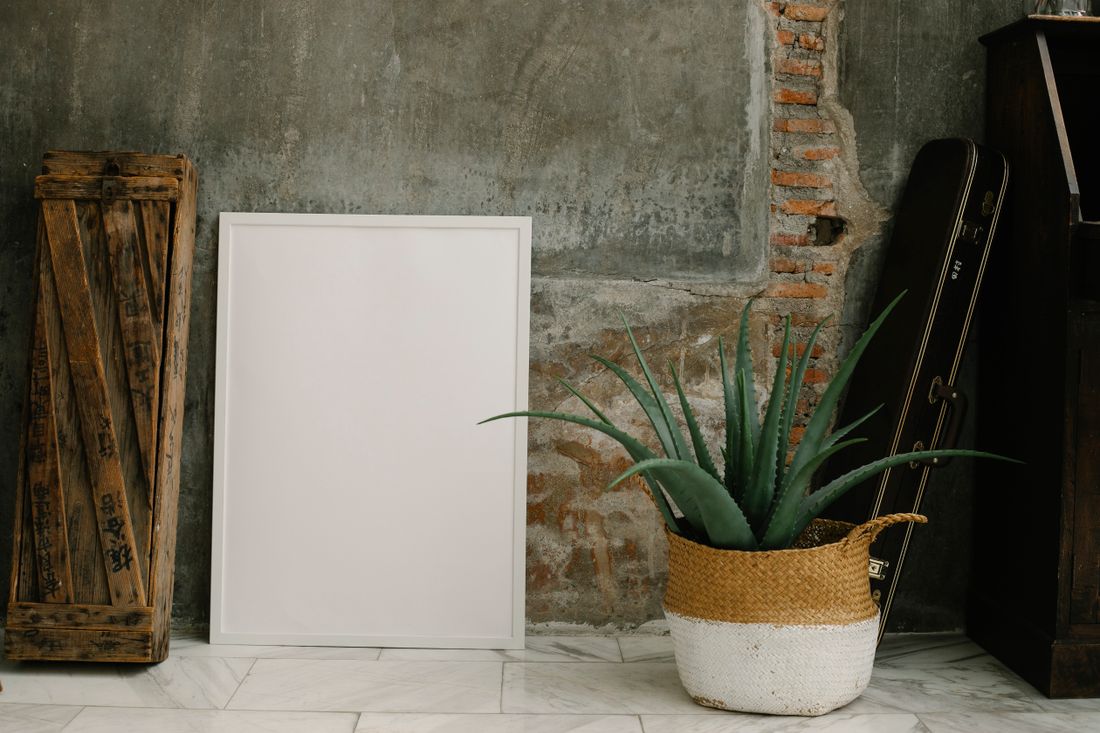 A blank framed piece leaning against a raw brick and concrete wall next to a wooden crate and a small potted plant